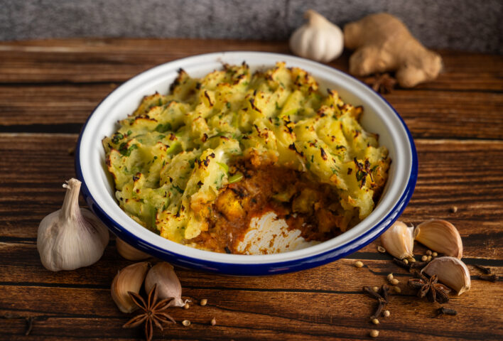 Goan curry inspired vegan fish pie in a white dish with golden mashed potato topping, garnished with herbs. A portion is removed to reveal the rich orange curry filling, surrounded by spices on a wooden surface